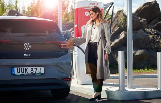 Woman in a long coat is charging a blue business vehicle at a Circle K station